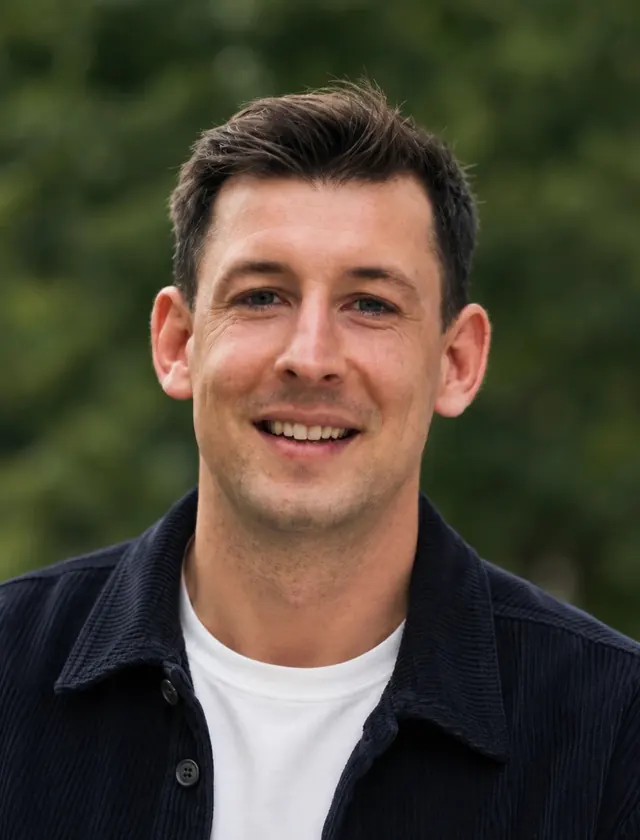 Tristan Kennedy lächelt, in dunkler Hemdjacke über weißem T-Shirt, im Hintergrund grüne Bäume